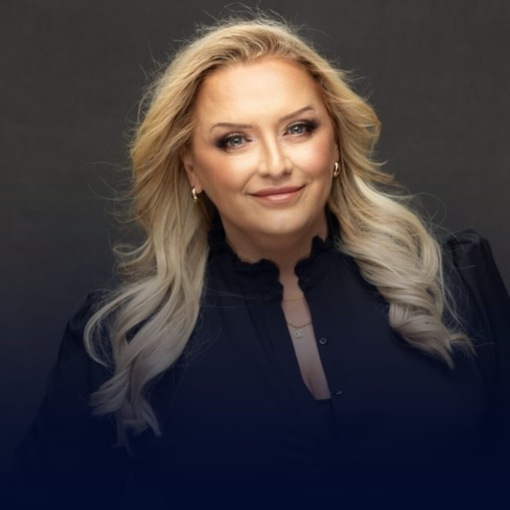 A close-up portrait of a smiling, middle-aged woman with long, wavy blonde hair wearing a dark jacket against a dark background.